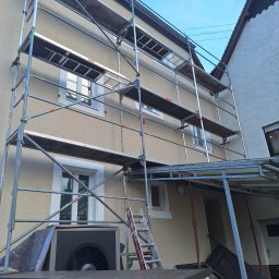 PerfectTynk - Elewacja domu w trakcie renowacji. Widoczne rusztowania, okna z białymi ramami i fragment sąsiedniego budynku. Na pierwszym planie jednostka zewnętrzna klimatyzacji.