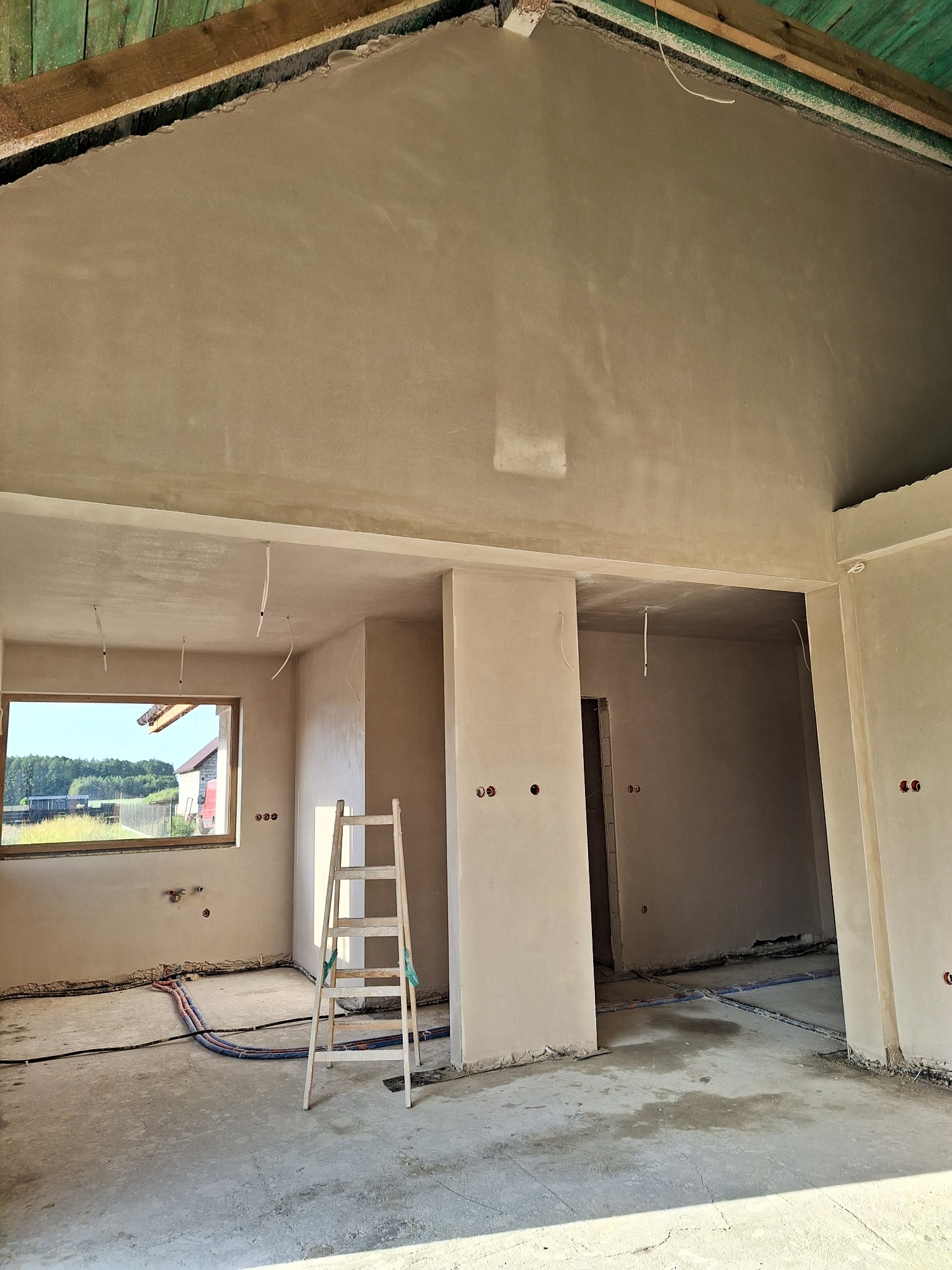 Wnętrze w stanie surowym zamkniętym po tynkowaniu, z drewnianą drabiną, widoczne instalacje elektryczne i hydrauliczne w podłodze, okno z widokiem na zewnątrz.