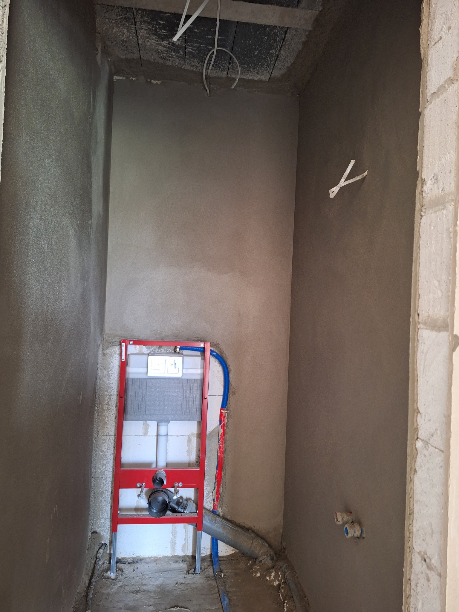 Ściany po nałożeniu tynku w łazience, widoczny stelaż podtynkowy WC z czerwonym profilem, elementy instalacji wodnej i kanalizacyjnej. Surowy stan wykończenia.