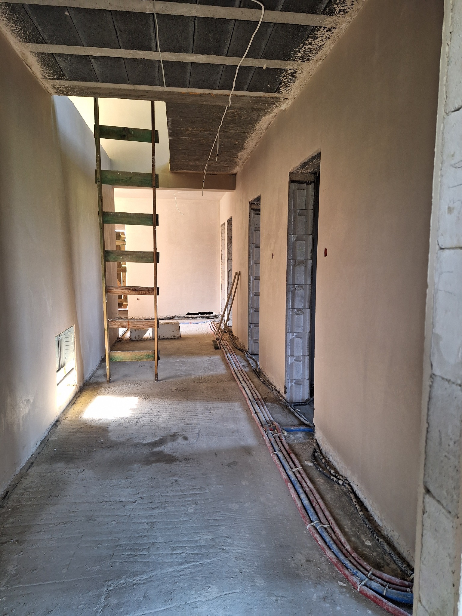Wnętrze w trakcie prac wykończeniowych: świeżo otynkowane ściany w beżowym odcieniu, drewniana drabina, instalacja wzdłuż ściany i surowy betonowy strop.