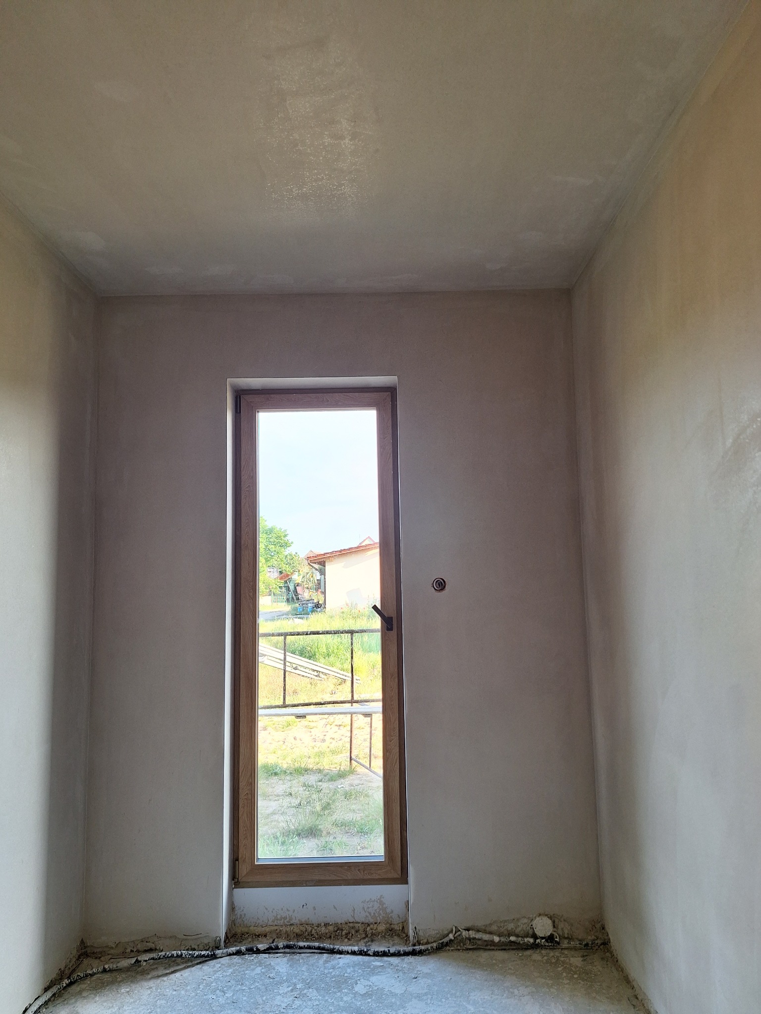 Wnętrze pokoju po wstępnym wyrównaniu ścian, widoczne szare tynki i drewniane okno balkonowe z widokiem na zewnątrz. Na podłodze widoczne instalacje.