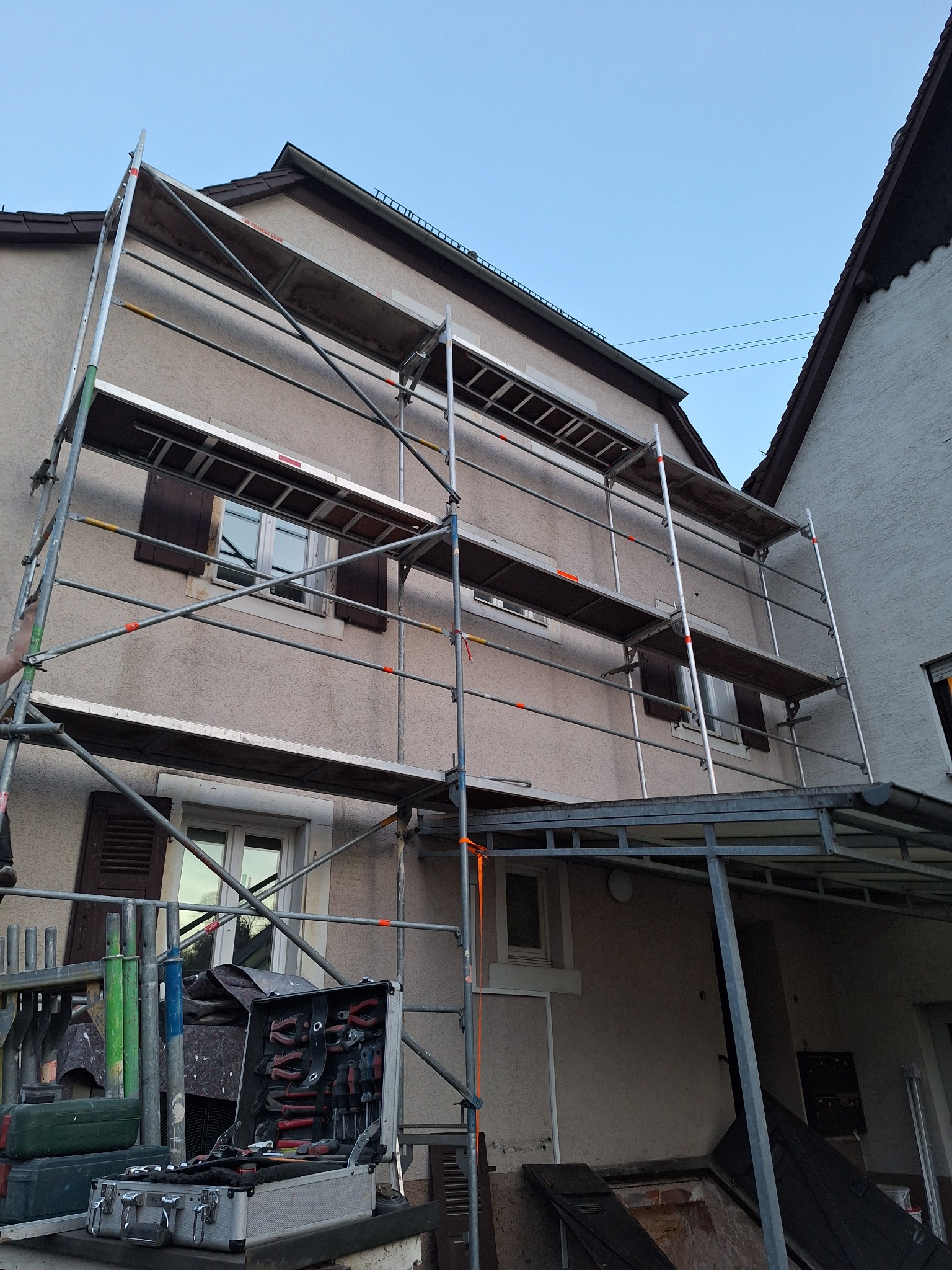 Elewacja domu w trakcie renowacji z użyciem rusztowania. Widoczna skrzynka z narzędziami i fragment drugiego budynku obok. Czyste niebo w tle.