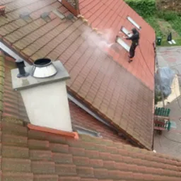 Pracownik w kombinezonie zabezpieczającym czyści dach z czerwonej dachówki za pomocą myjki ciśnieniowej. Widoczna różnica między umytą a zabrudzoną częścią dachu.