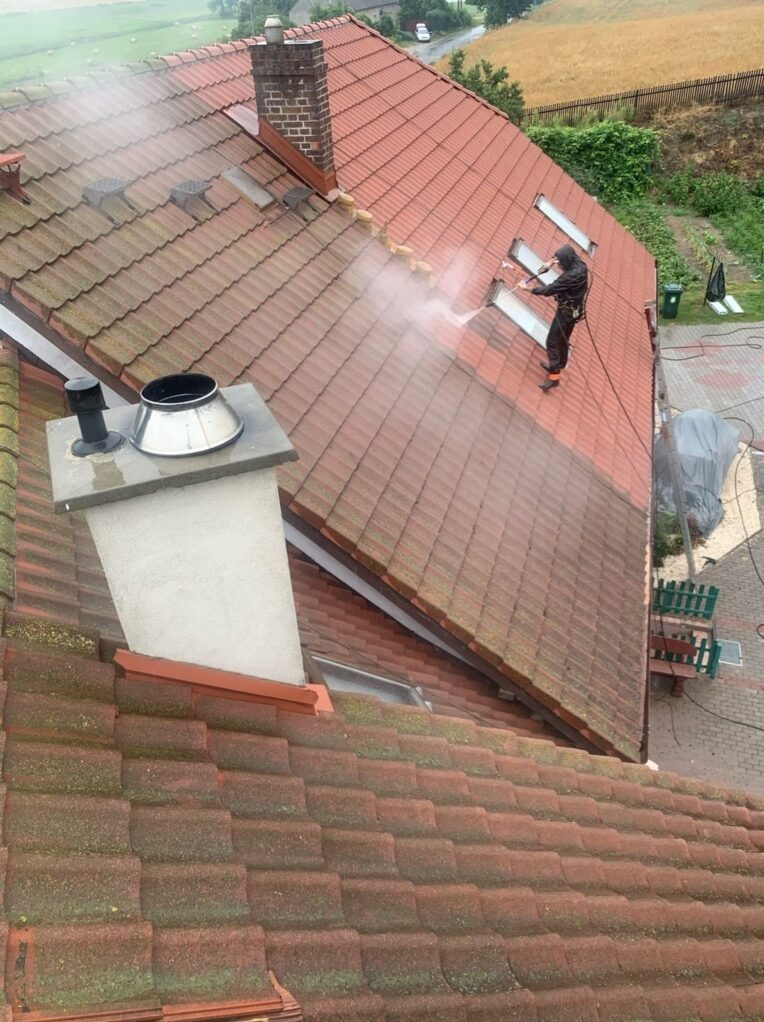Pracownik w kombinezonie zabezpieczającym czyści dach z czerwonej dachówki za pomocą myjki ciśnieniowej. Widoczna różnica między umytą a zabrudzoną częścią dachu.