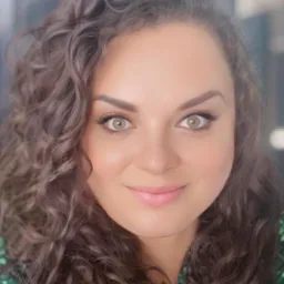 Portret kobiety o kręconych włosach i jasnych oczach, z delikatnym uśmiechem, na rozmytym tle miejskim. Ubrana w zieloną bluzkę w kwiaty.