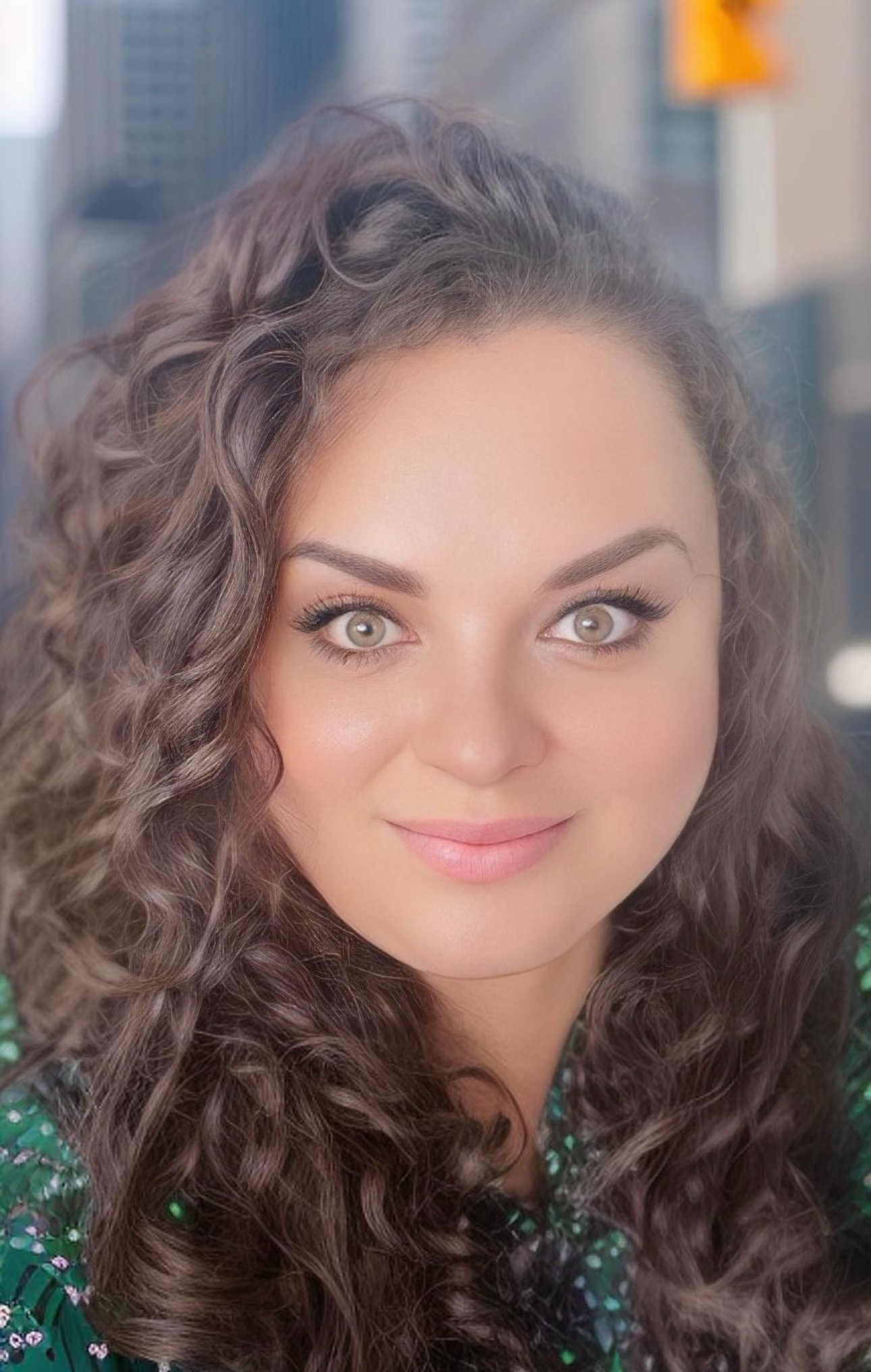 Portret kobiety o kręconych włosach i jasnych oczach, z delikatnym uśmiechem, na rozmytym tle miejskim. Ubrana w zieloną bluzkę w kwiaty.