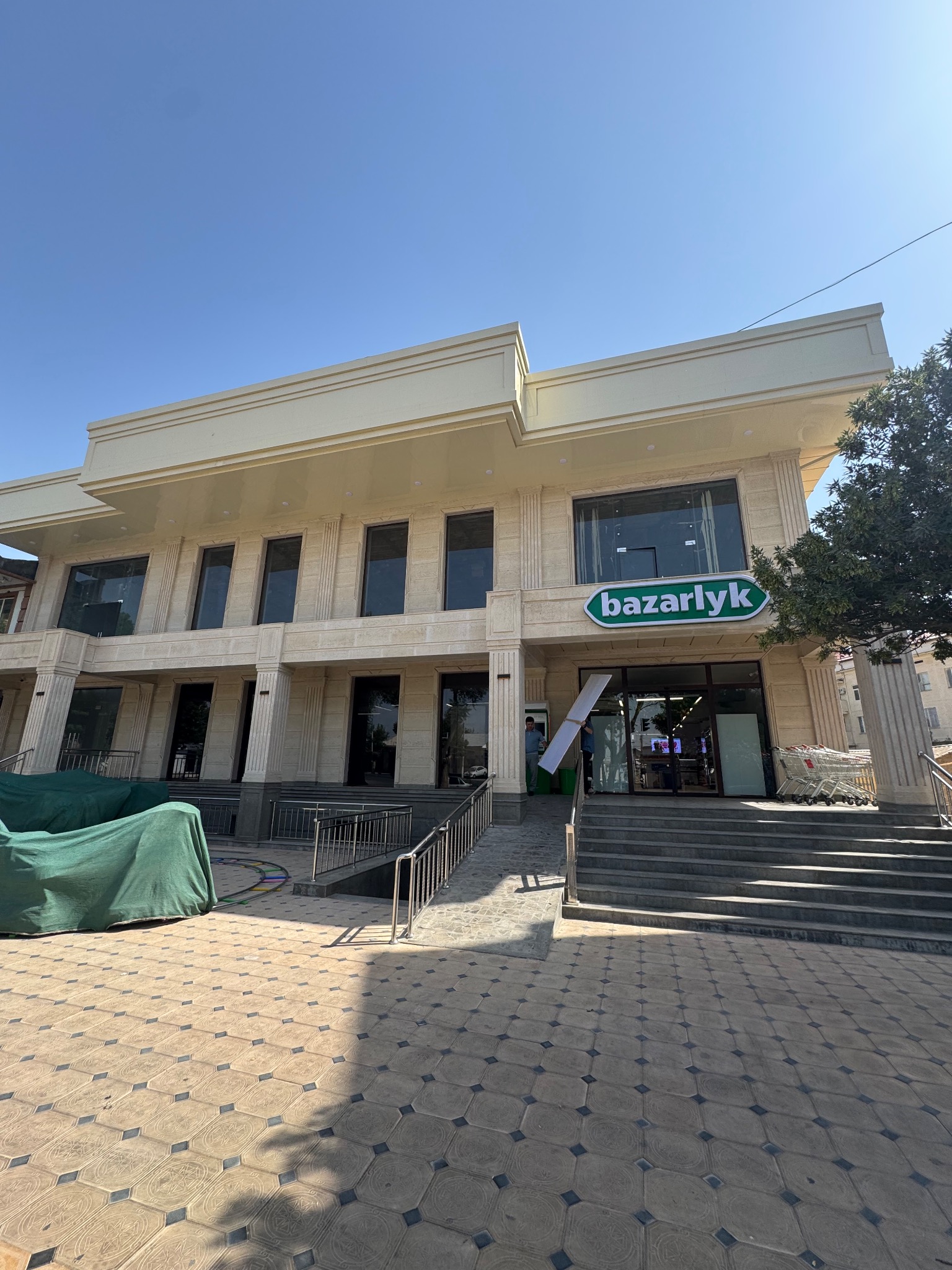 Dwupiętrowy budynek z beżową elewacją i szyldem 'bazarlyk' nad wejściem. Widoczne schody, balustrady i chodnik z wzorzystej kostki. Część budynku zakryta zieloną plandeką.
