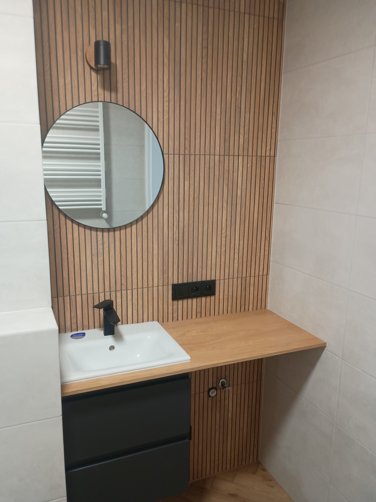 Nowoczesna łazienka z umywalką nablatową, czarną armaturą i okrągłym lustrem na tle ściany wykończonej drewnianymi lamelami. Widoczne gniazdko elektryczne.