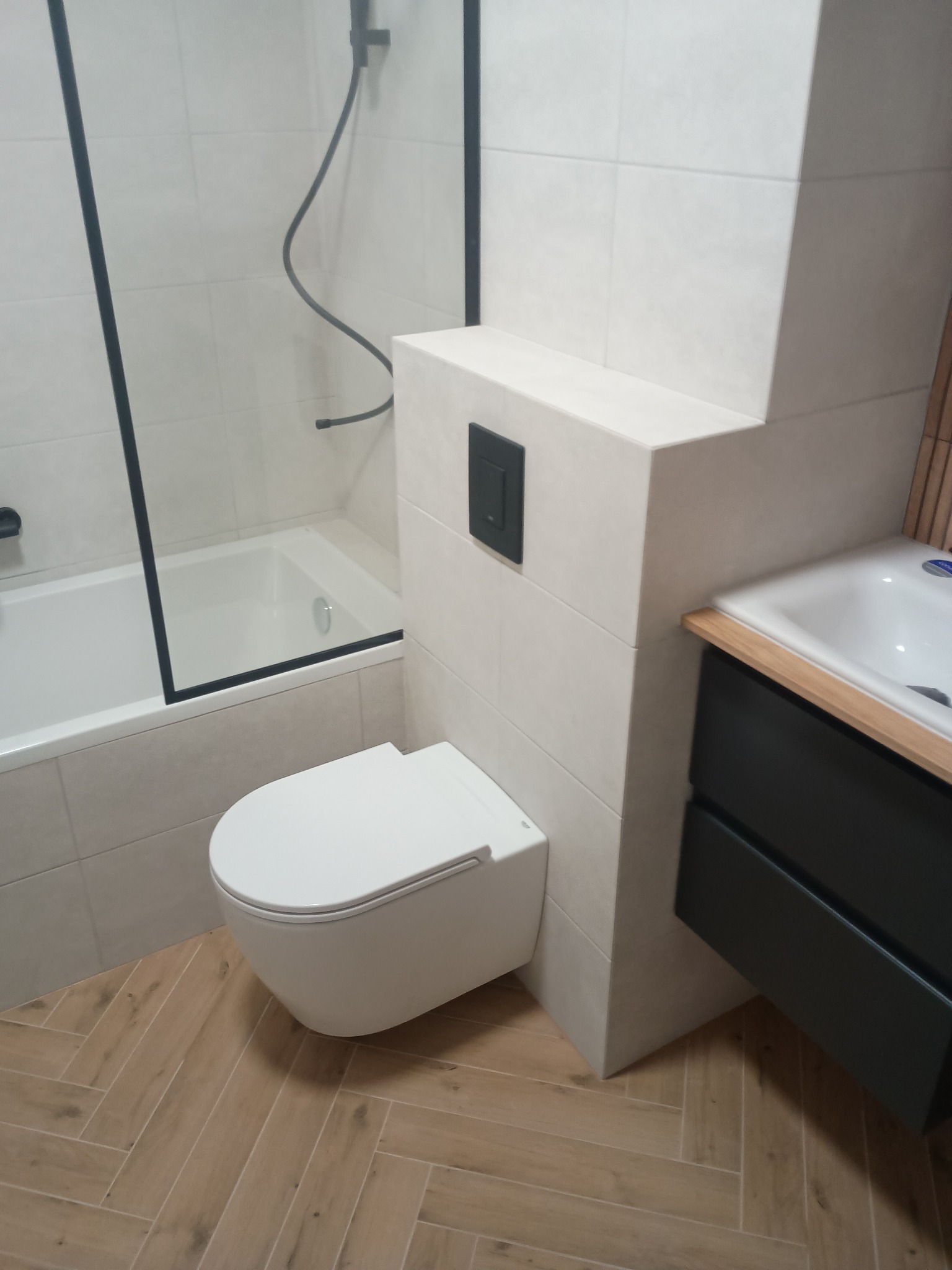 Nowoczesna łazienka z podwieszaną toaletą, kabiną prysznicową z czarną ramą i szafką umywalkową. Parkiet ułożony w jodełkę. Minimalistyczny design z akcentami czerni.