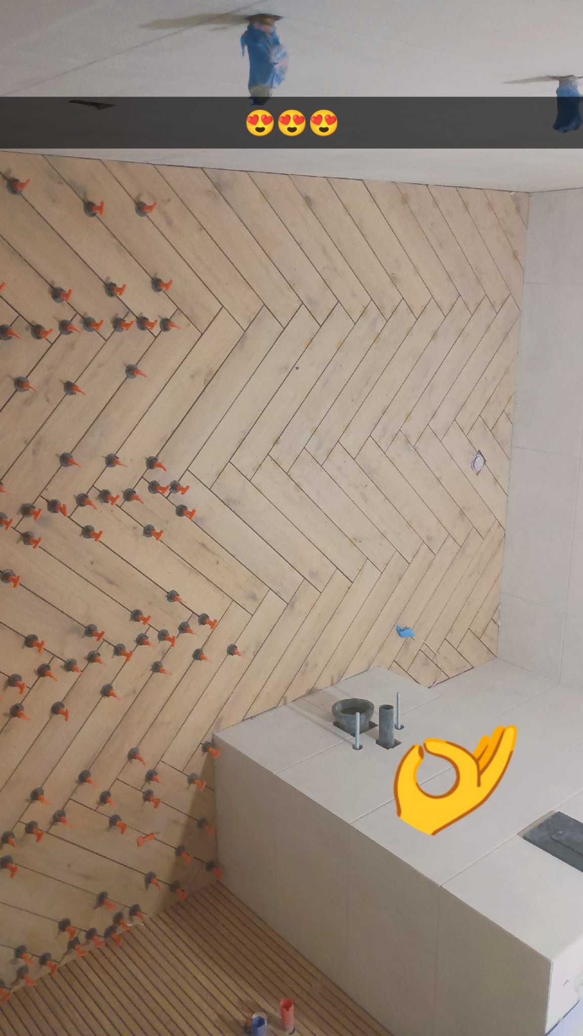 Ściana wyłożona jasnymi płytkami w jodełkę, z pomarańczowymi krzyżykami dystansowymi. Widoczny fragment wanny w trakcie wykańczania. Na suficie zabezpieczone lampy.