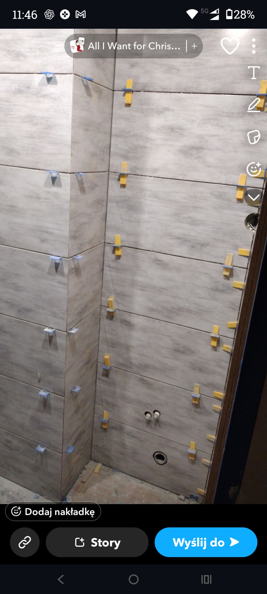 Ściana w trakcie układania płytek ceramicznych w łazience. Widoczne krzyżyki dystansowe i otwory na instalacje. Płytki w odcieniach szarości z delikatnym wzorem.