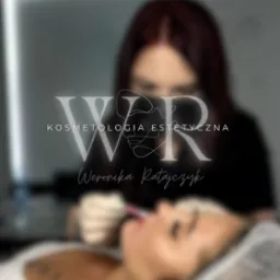 Zamazane ujęcie z zabiegu na usta z logotypem firmy. Igła w ustach klientki. Widoczna twarz osoby wykonującej zabieg. Stonowana kolorystyka i rozmyte tło.