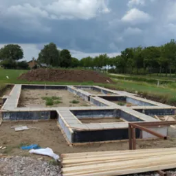 Zalane betonem szalunki fundamentowe domu jednorodzinnego, widok z góry. Na pierwszym planie stos drewna, w tle kopiec ziemi i dom w oddali.