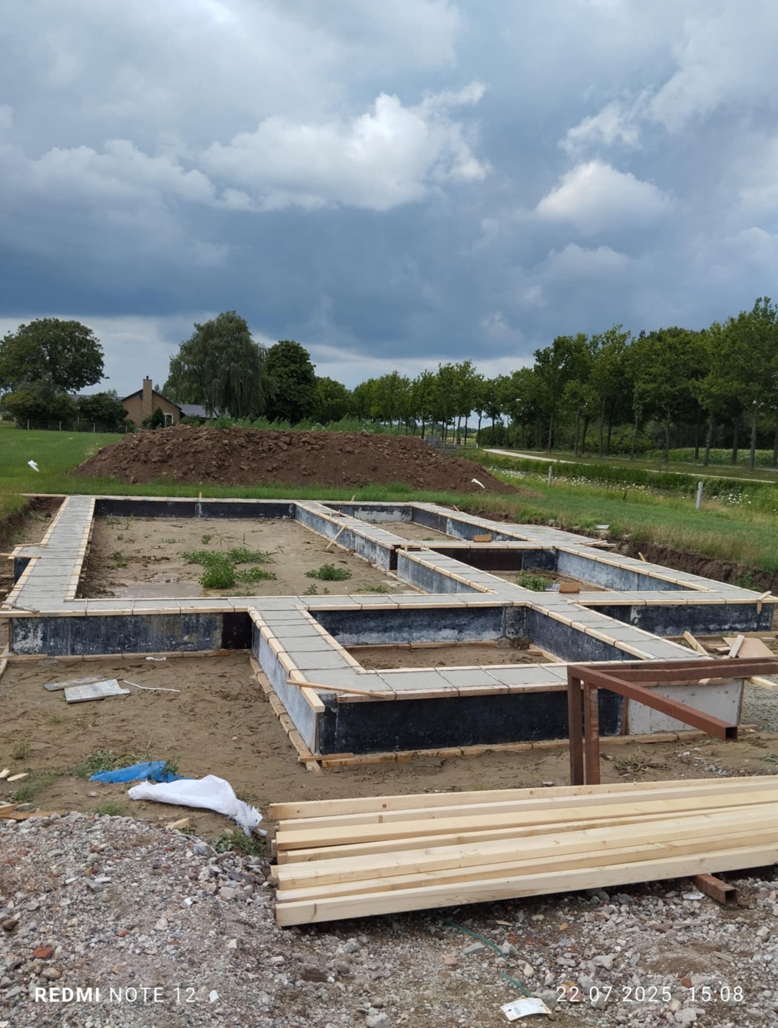 Zalane betonem szalunki fundamentowe domu jednorodzinnego, widok z góry. Na pierwszym planie stos drewna, w tle kopiec ziemi i dom w oddali.