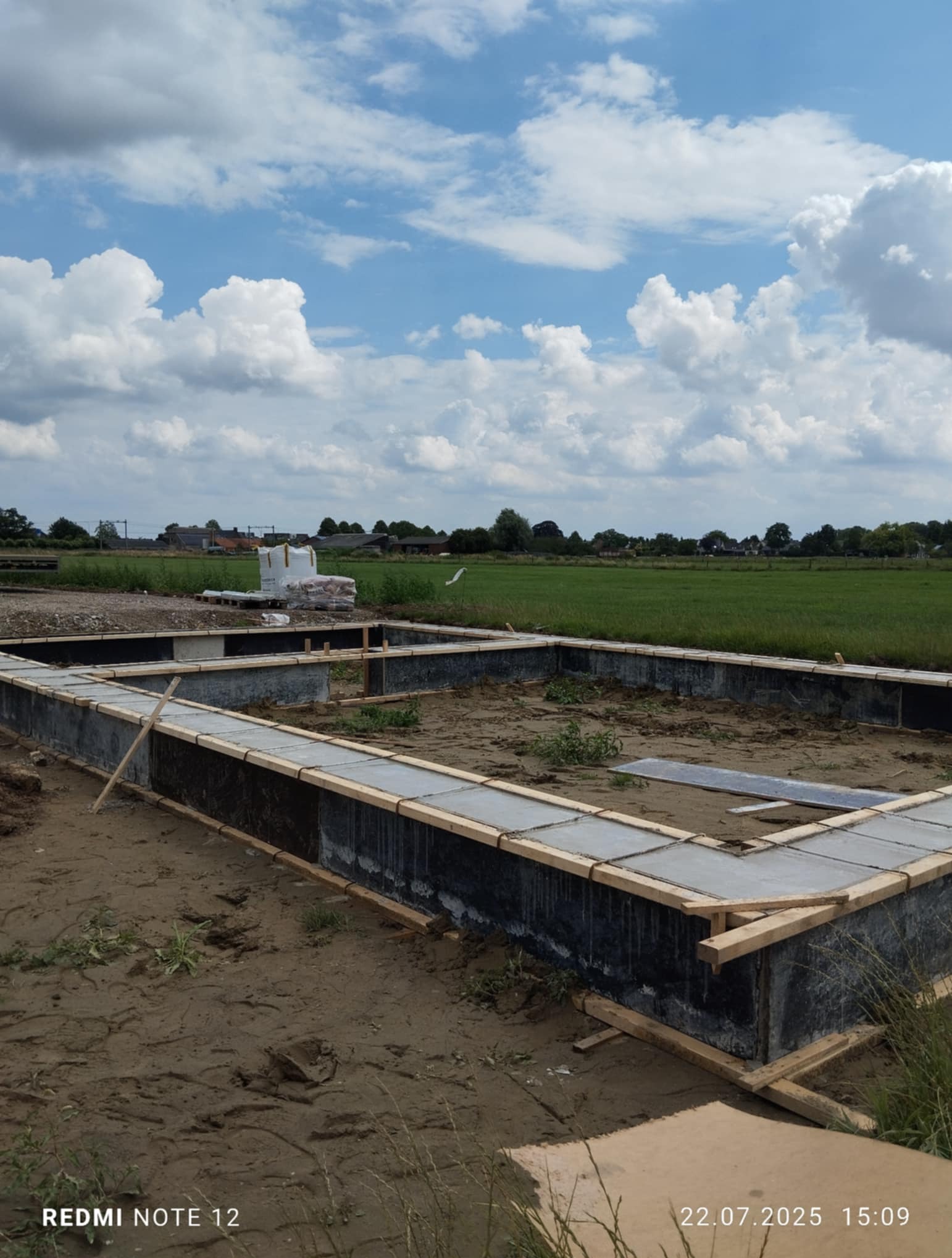 Zalane betonem szalunki fundamentowe z drewnianym oparciem, na tle zielonego pola i błękitnego nieba z chmurami. Widoczna izolacja pionowa. Prace budowlane w toku.