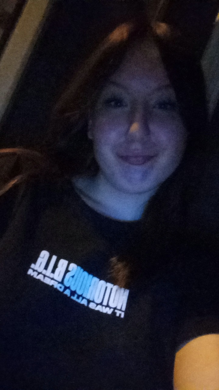 Portret kobiety w ciemnym otoczeniu, uśmiechniętej, z napisem na koszulce. Słabe oświetlenie, widoczne detale twarzy i fragment koszulki.