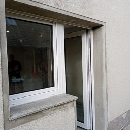 Figury ogrodowe montaż drzwi - Nowe, białe drzwi przesuwne na zewnątrz budynku z szarą elewacją. Widoczne ślady świeżego tynku wokół ościeżnicy. W tle odbicie sylwetki mężczyzny w szybie.