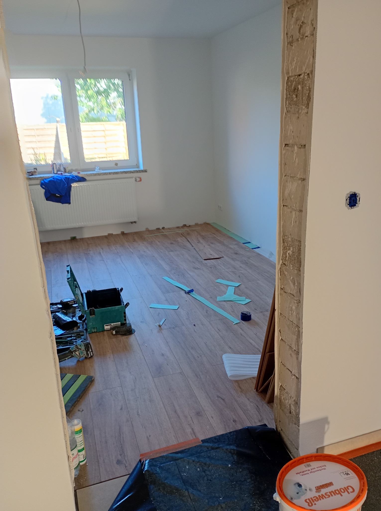 Pokój w trakcie układania paneli podłogowych. Widoczne narzędzia, klej i fragmenty paneli. Częściowo ułożona podłoga w odcieniach brązu. Okno w tle.