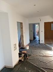 Wnętrze w trakcie remontu: zabezpieczona podłoga, surowe ściany, otwór drzwiowy z widoczną konstrukcją. Etap przygotowawczy do wykończenia.