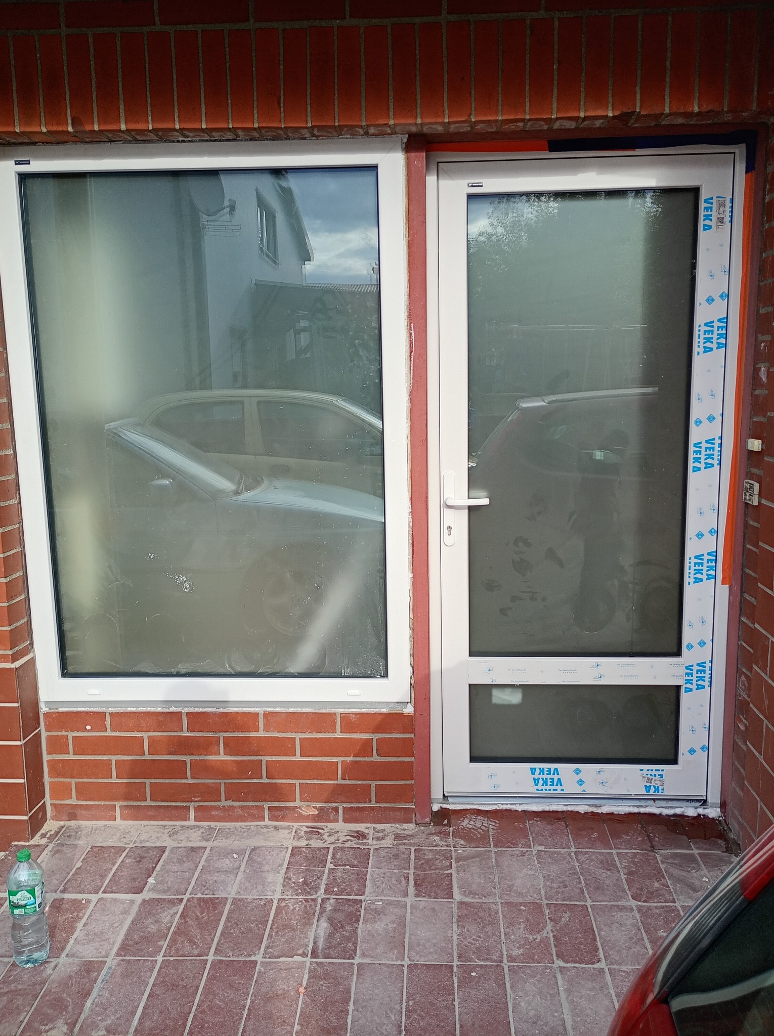 Nowe, białe drzwi i okno PCV zamontowane w czerwonej, ceglanej ścianie budynku. Widoczne folie ochronne na profilach VEKA.