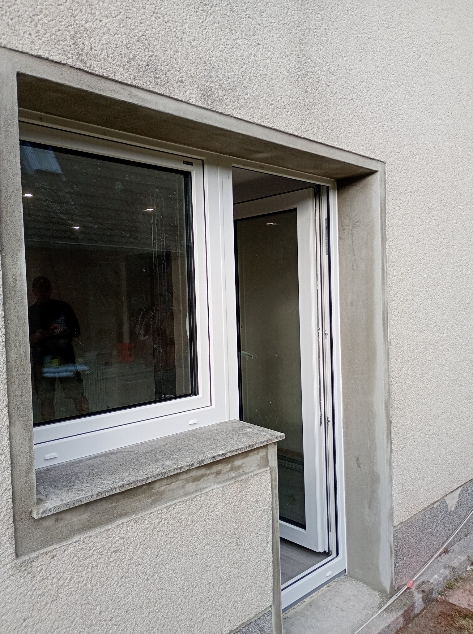 Nowe, białe drzwi przesuwne na zewnątrz budynku z szarą elewacją. Widoczne ślady świeżego tynku wokół ościeżnicy. W tle odbicie sylwetki mężczyzny w szybie.