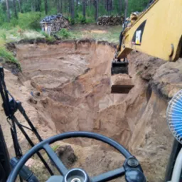 Żółta koparka CAT nabiera ziemię w wykopie, widok z kabiny operatora na tle lasu. Widoczne warstwy gruntu w ścianach wykopu i sterta drewna w tle.