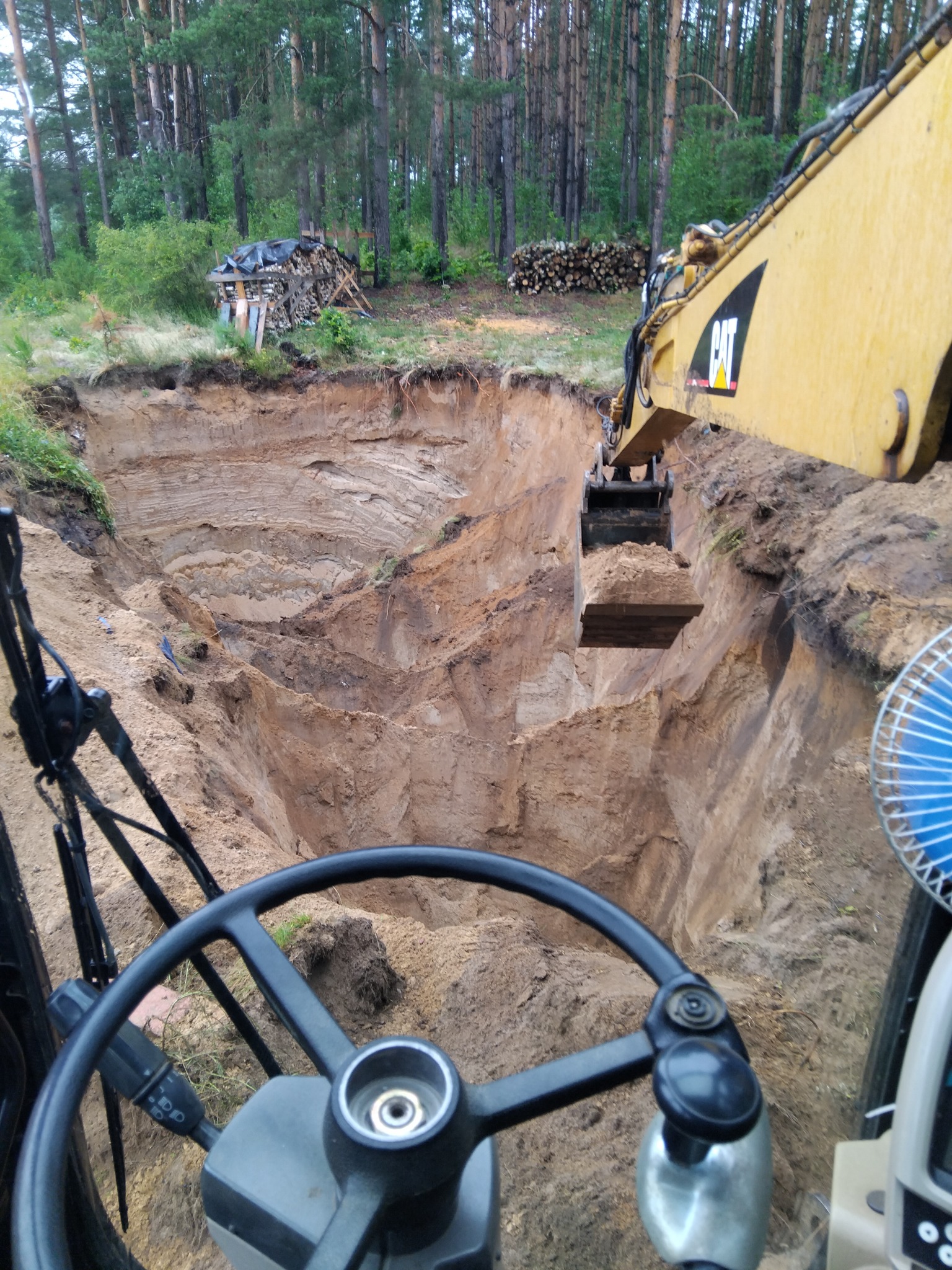 Żółta koparka CAT nabiera ziemię w wykopie, widok z kabiny operatora na tle lasu. Widoczne warstwy gruntu w ścianach wykopu i sterta drewna w tle.
