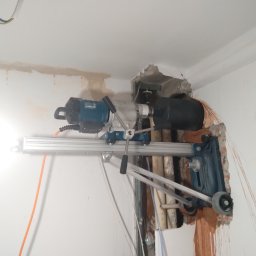 HVAC Instalacje Robert Knotek - Wiertnica diamentowa w trakcie wiercenia otworu w ścianie, widoczne przewody i rury. Praca w toku, pył i fragmenty muru wokół otworu. Specjalistyczny sprzęt do precyzyjnych prac instalacyjnych.