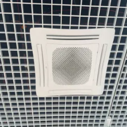 Biała, kwadratowa klimatyzacja kasetonowa wkomponowana w sufit rastrowy z widocznymi przewodami. Oświetlenie punktowe na szynie.