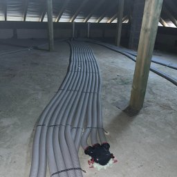 HVAC Instalacje Robert Knotek - Montaż rekuperacji na poddaszu: szare rury wentylacyjne ułożone równolegle, przymocowane opaskami, widoczny rozdzielacz i drewniana konstrukcja dachu.