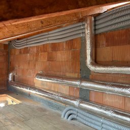 HVAC Instalacje Robert Knotek - Montaż rekuperacji: rury wentylacyjne, część owinięta folią izolacyjną, prowadzone wzdłuż ceglanej ściany i drewnianego stropu w surowym stanie budynku.