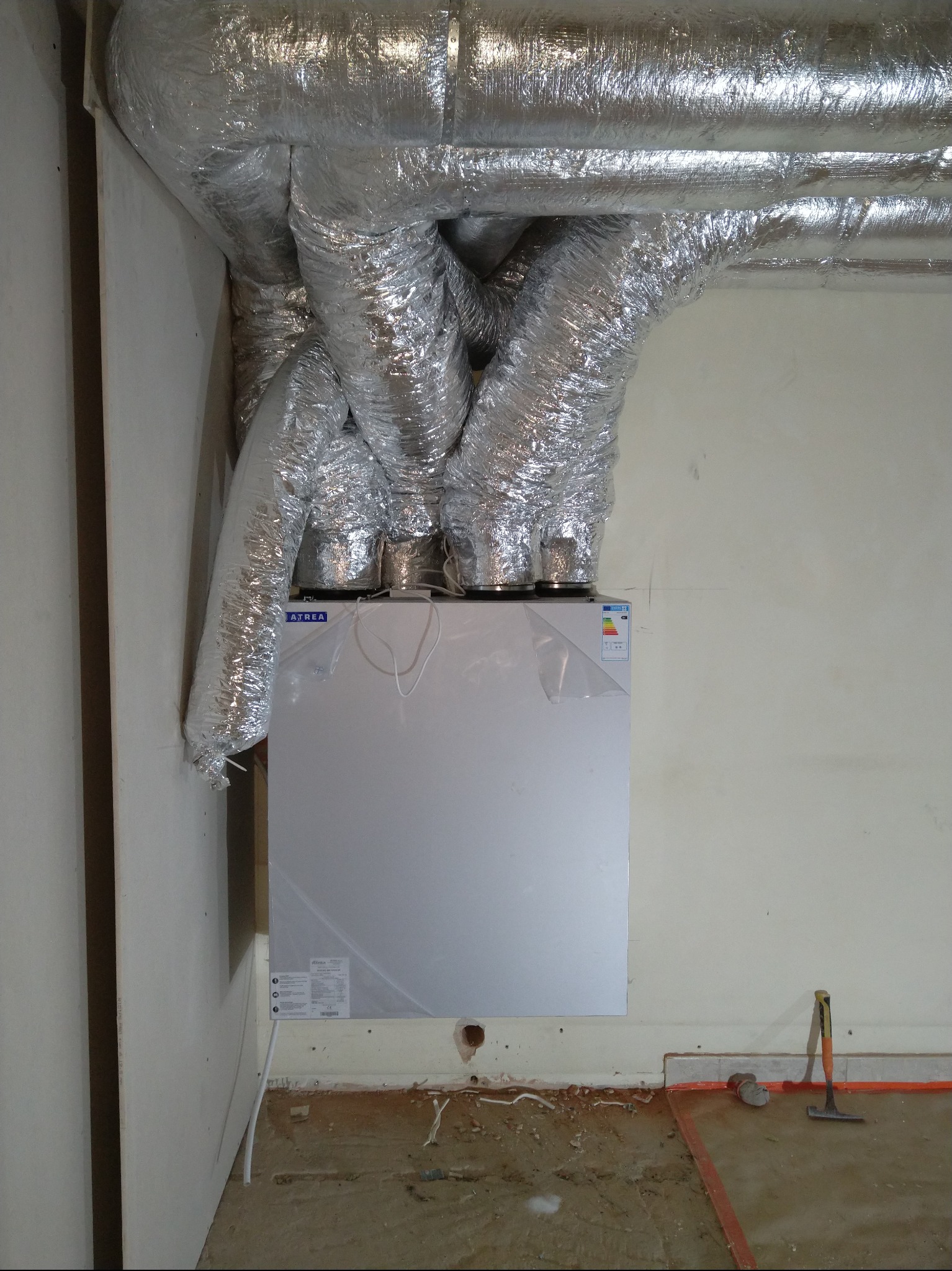 Centrala rekuperacyjna Atrea z podłączonymi przewodami wentylacyjnymi w folii aluminiowej, zamontowana na ścianie w surowym pomieszczeniu. Widoczne narzędzia.