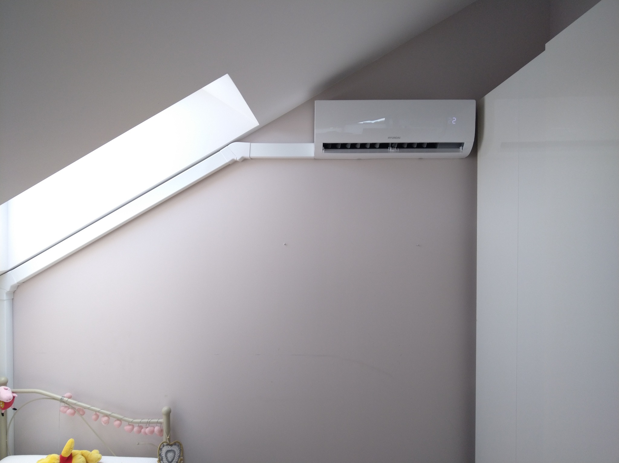Biały klimatyzator Hyundai na ścianie pokoju dziecięcego z oknem dachowym. Widoczne rury instalacyjne i fragment łóżka z zabawkami. Jasne, naturalne oświetlenie.
