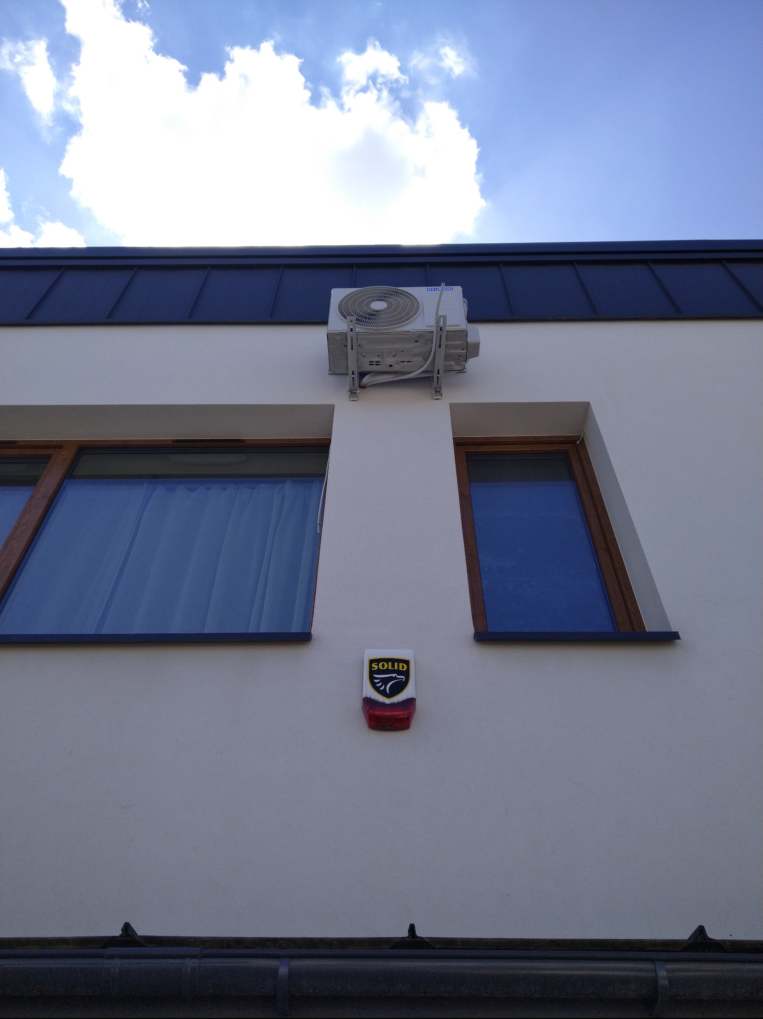 Biała elewacja domu z zamontowaną klimatyzacją Hyundai, widoczna między dwoma oknami. Alarm Solid pod oknami, niebieskie niebo z chmurami w tle.