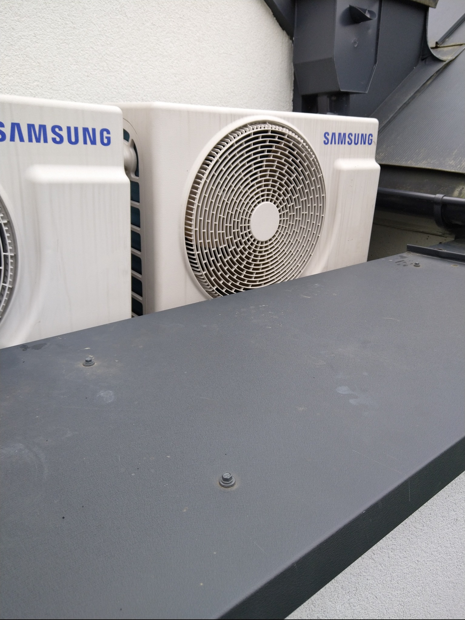 Dwa białe, zewnętrzne moduły klimatyzacji Samsung na tle jasnej ściany i szarego dachu, widoczne z bliska pod kątem, z szarą metalową powierzchnią na pierwszym planie.