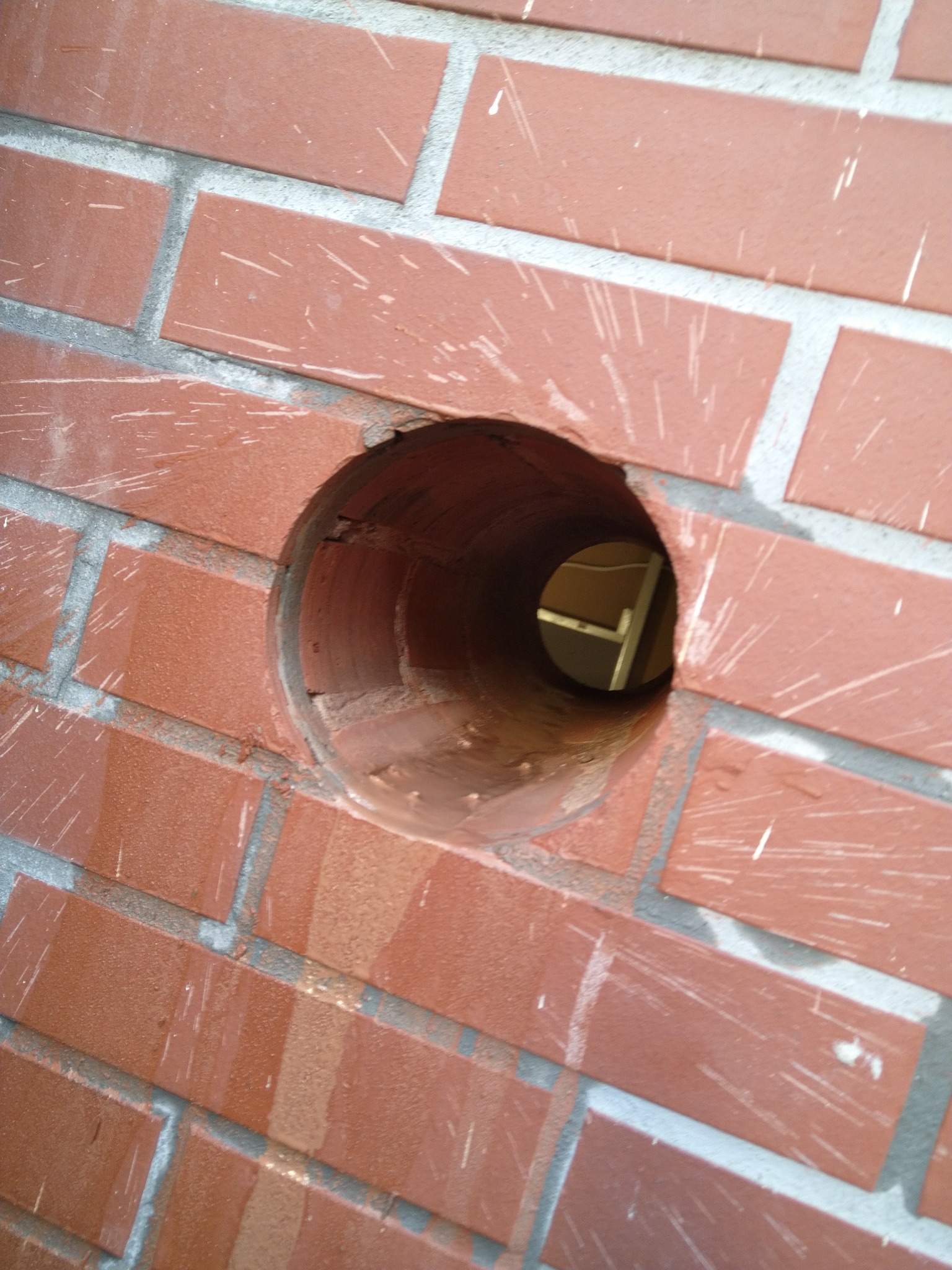 Otwór w ceglanej ścianie, prawdopodobnie przygotowany pod montaż instalacji wentylacyjnej lub klimatyzacyjnej; widoczne ślady wiercenia.