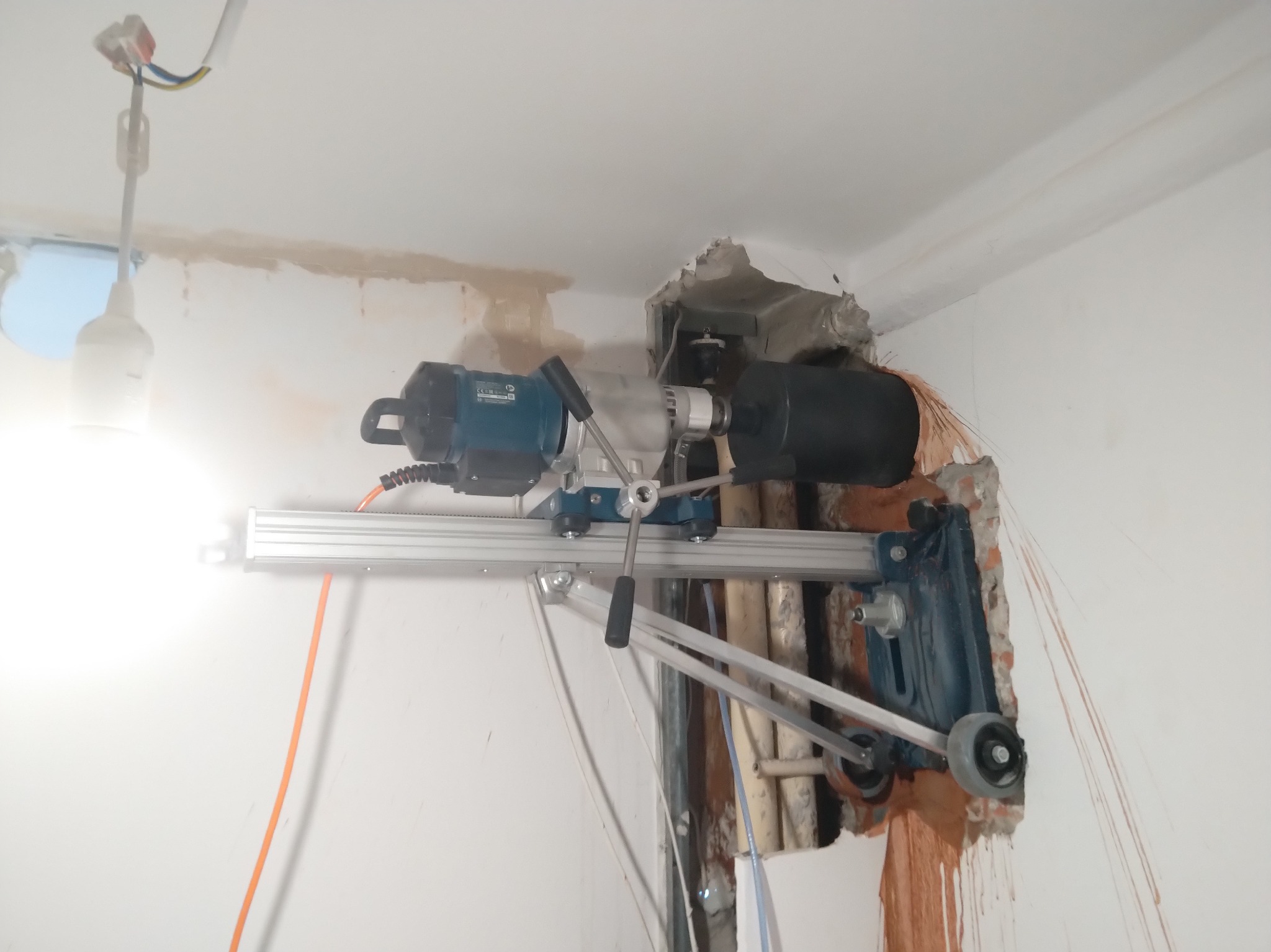 Wiertnica diamentowa w trakcie wiercenia otworu w ścianie, widoczne przewody i rury. Praca w toku, pył i fragmenty muru wokół otworu. Specjalistyczny sprzęt do precyzyjnych prac instalacyjnych.