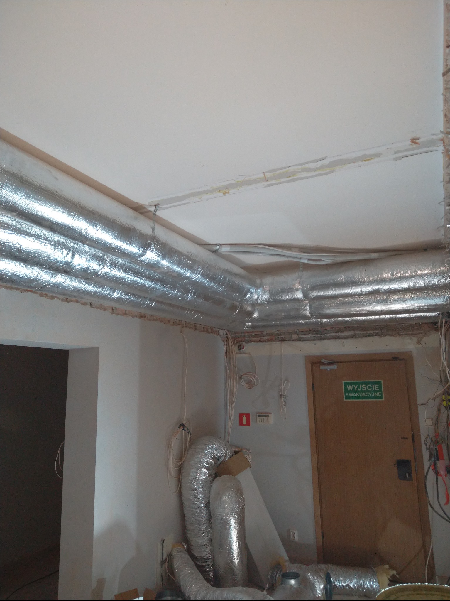 Montaż srebrnych, izolowanych przewodów wentylacyjnych pod sufitem w trakcie prac wykończeniowych. Widoczne fragmenty instalacji i drzwi ewakuacyjne.