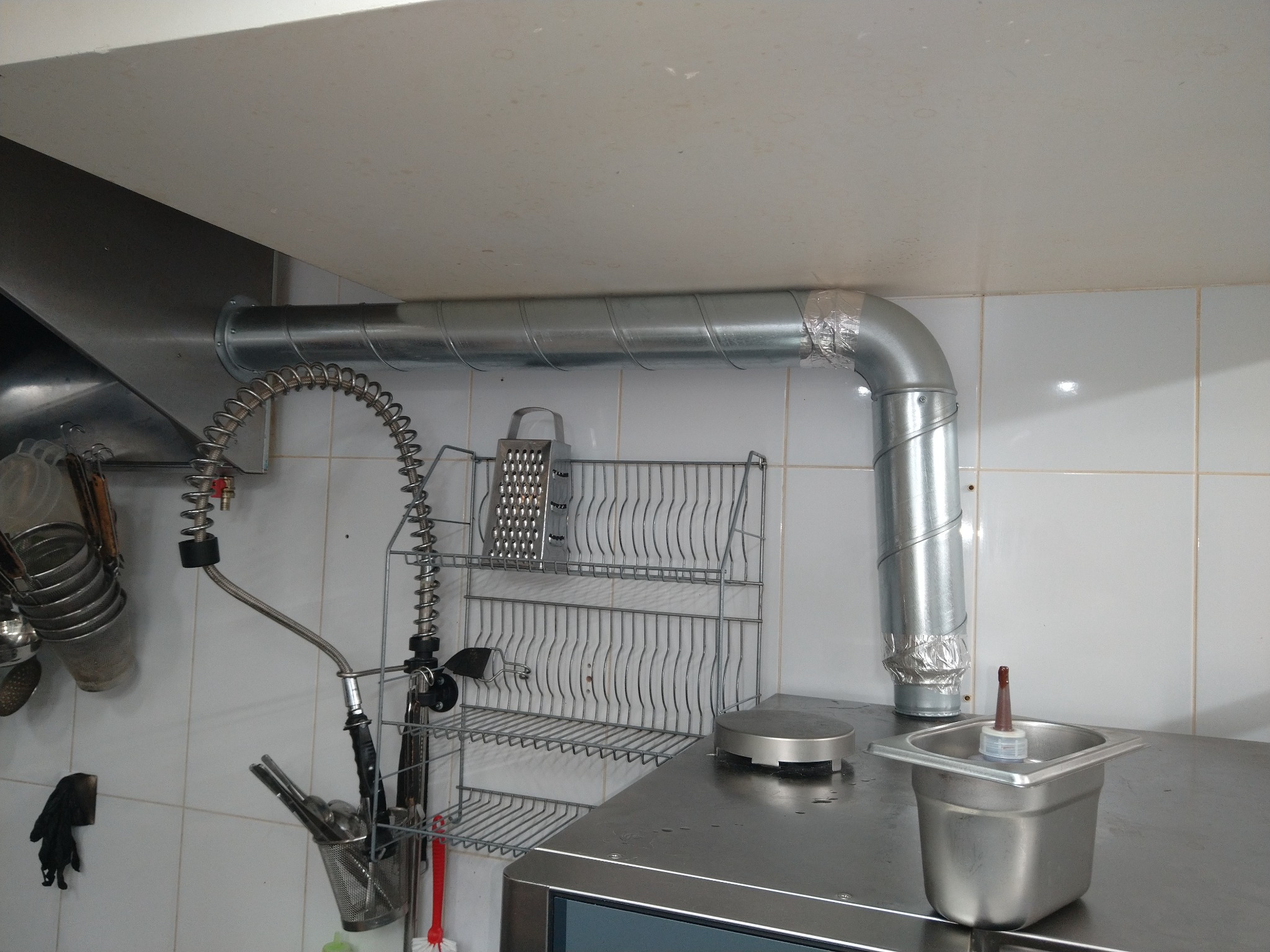 Metalowy kanał wentylacyjny, spiralny, przymocowany do okapu w kuchni, zabezpieczony taśmą. Widoczne akcesoria kuchenne i białe płytki na ścianie.