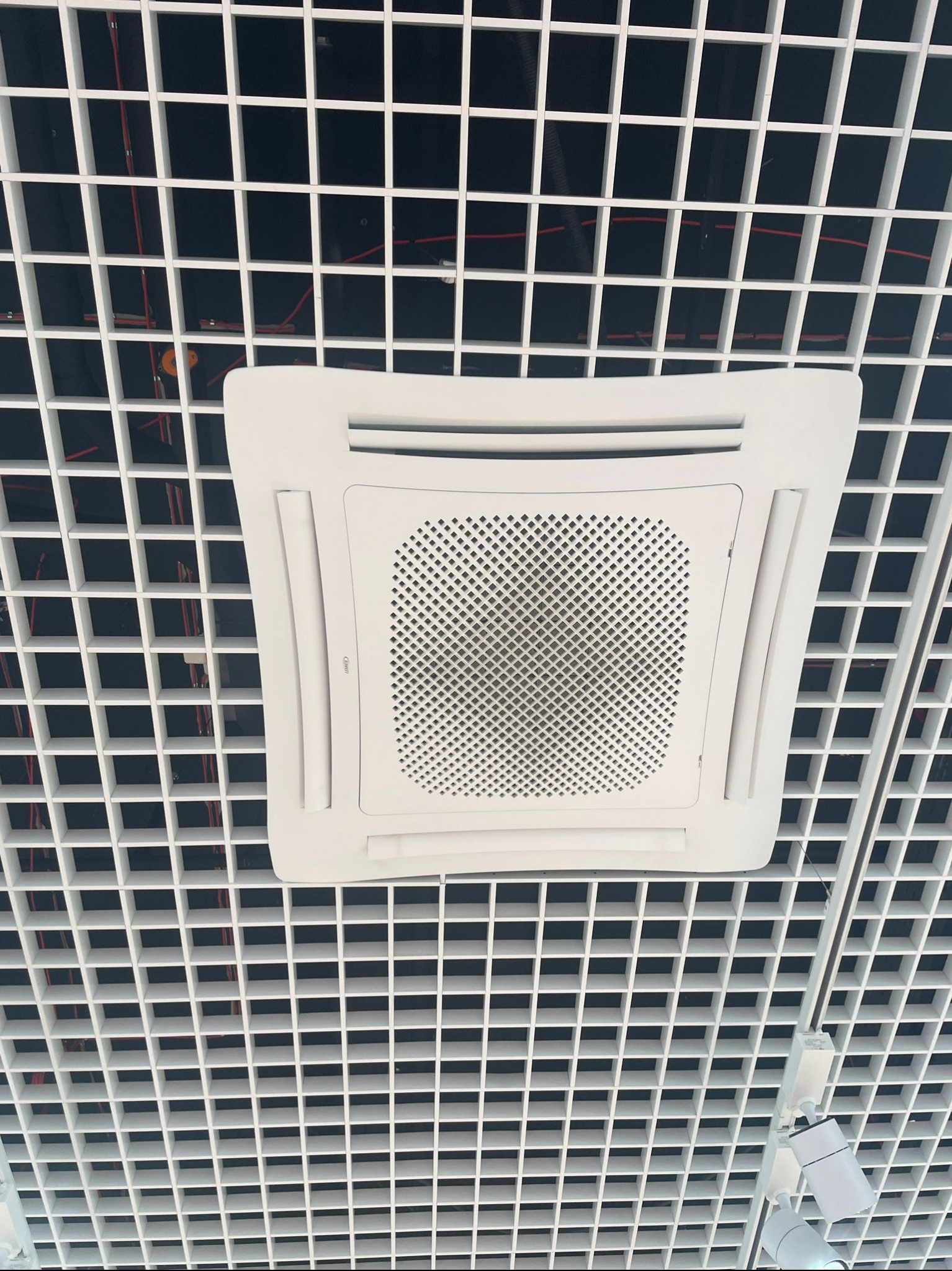 Biała, kwadratowa klimatyzacja kasetonowa wkomponowana w sufit rastrowy z widocznymi przewodami. Oświetlenie punktowe na szynie.