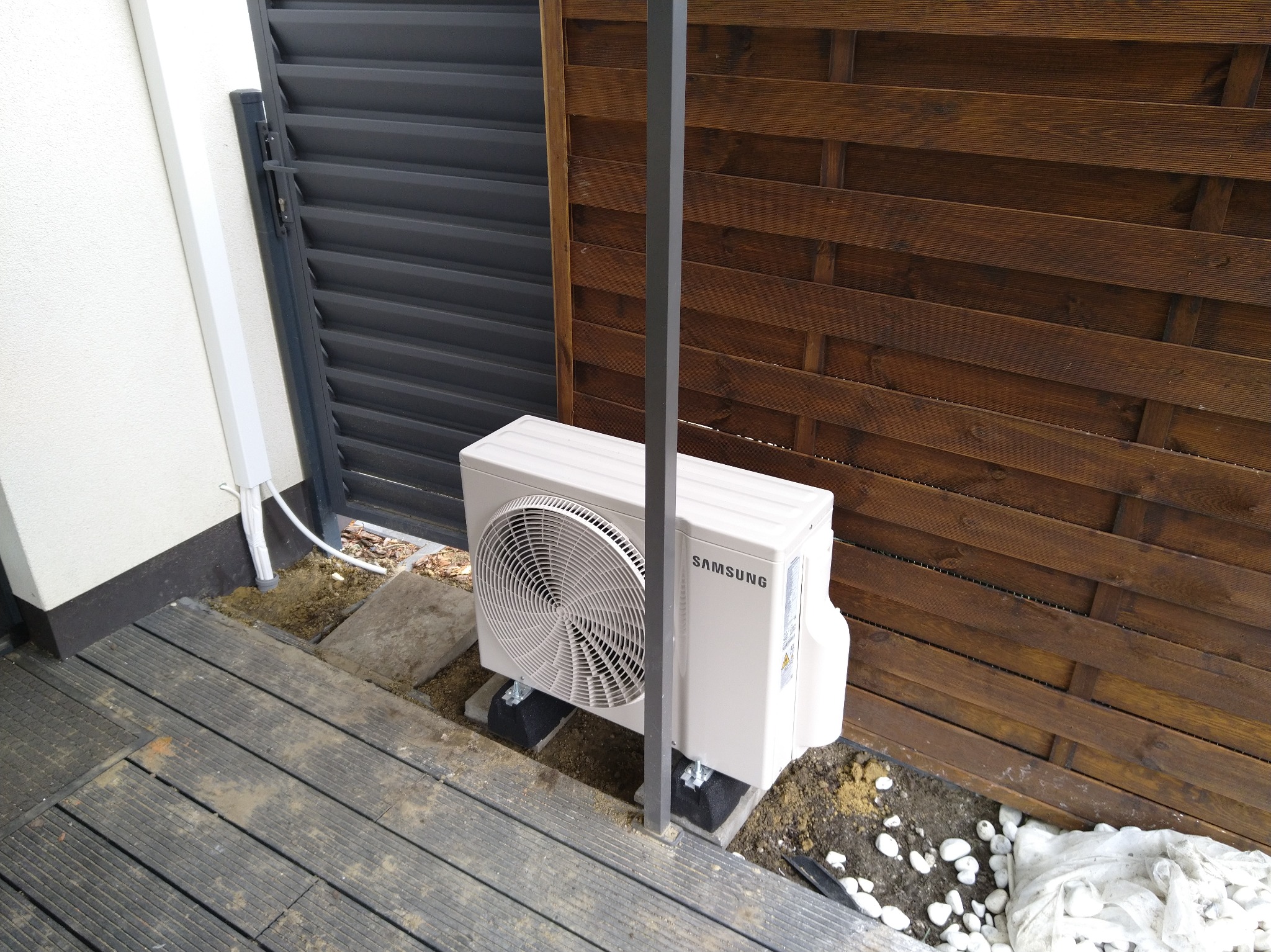 Biała jednostka zewnętrzna klimatyzacji Samsung, zamontowana na betonowych podstawach przy drewnianym płocie i szarej bramie, widok z boku.