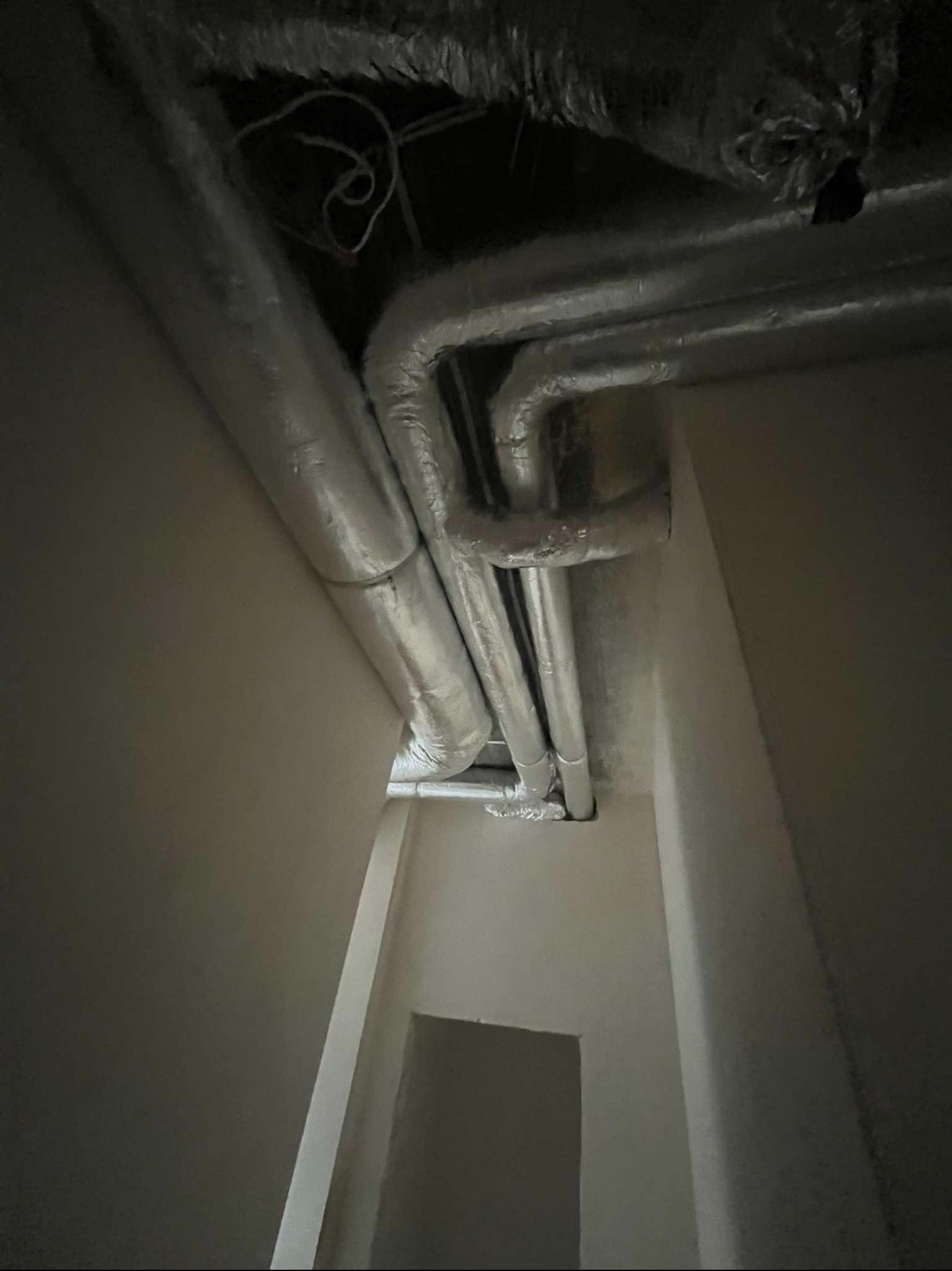 Srebrne rury wentylacyjne pod sufitem, widoczne przewody i instalacja, kadr z dołu, szare ściany, w tle otwór drzwiowy.