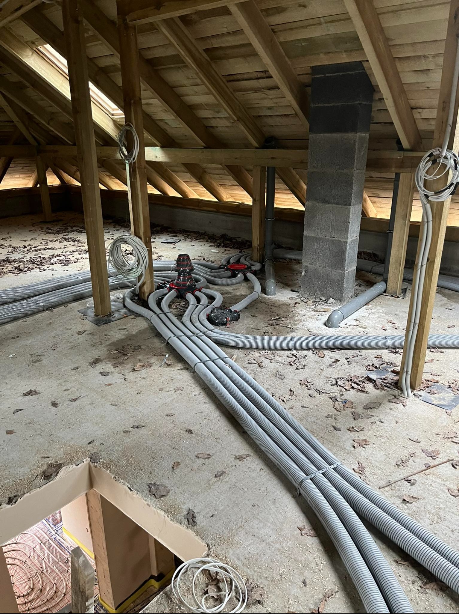 Szare rury wentylacyjne prowadzone na poddaszu z drewnianą konstrukcją i kominem z pustaków. Widoczny system rozdzielczy i fragment instalacji ogrzewania podłogowego.