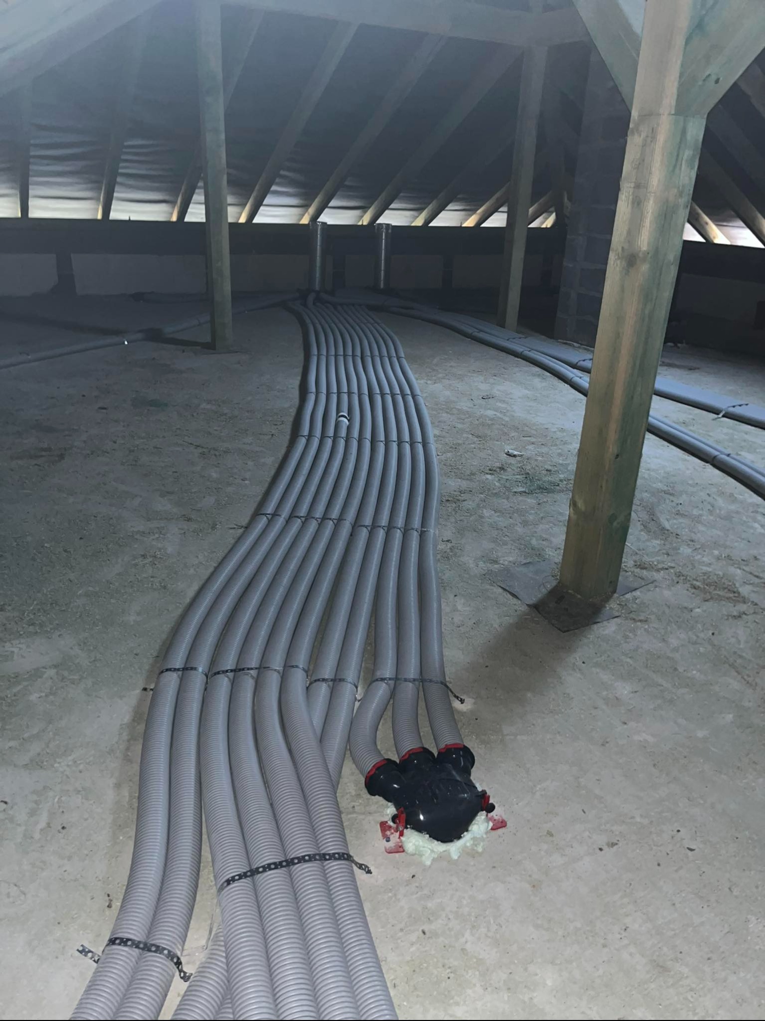 Montaż rekuperacji na poddaszu: szare rury wentylacyjne ułożone równolegle, przymocowane opaskami, widoczny rozdzielacz i drewniana konstrukcja dachu.