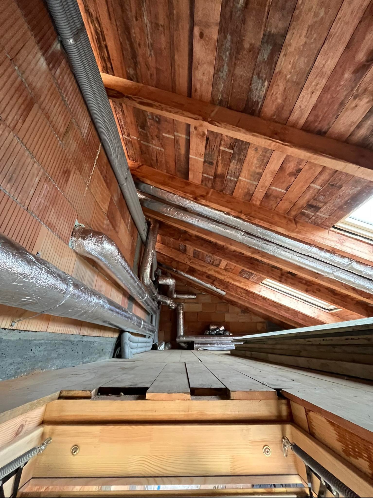 Instalacja wentylacyjna na poddaszu z drewnianym stropem i ceglaną ścianą. Widoczne rury wentylacyjne w izolacji aluminiowej i szare rury karbowane.