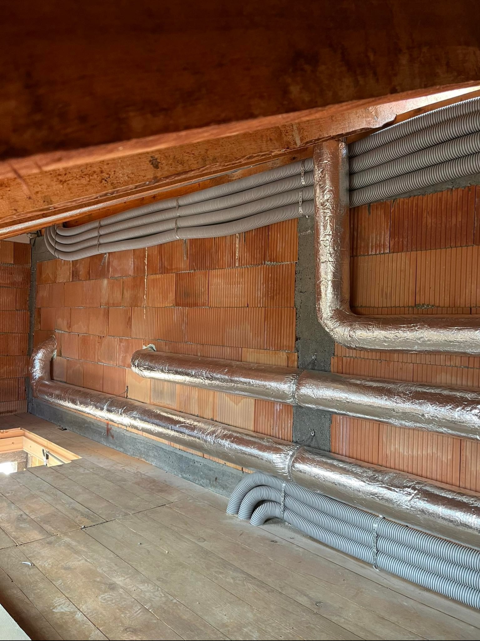 Montaż rekuperacji: rury wentylacyjne, część owinięta folią izolacyjną, prowadzone wzdłuż ceglanej ściany i drewnianego stropu w surowym stanie budynku.