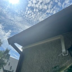 Arktus Klimatyzacja Piotr Respondek - Biała rura instalacji klimatyzacyjnej zamontowana na teksturowanej ścianie budynku, widoczna od dołu na tle błękitnego nieba z chmurami i słońcem.