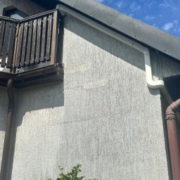 Arktus Klimatyzacja Piotr Respondek - Fragment elewacji budynku z drewnianą balustradą balkonu, brązową rynną i białym kanałem instalacyjnym klimatyzacji na tle błękitnego nieba.