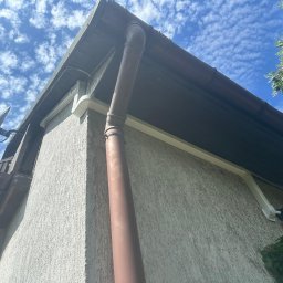 Arktus Klimatyzacja Piotr Respondek - Widok z dołu na narożnik budynku z beżową elewacją, brązową rynną i rurą spustową, oraz białym korytem instalacji klimatyzacji, częściowo widoczną jednostką zewnętrzną i anteną satelitarną na tle...