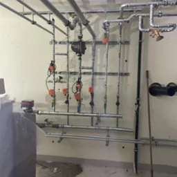 Złożona instalacja grzewcza z rurami, zaworami i pompą obiegową. Widoczne elementy mocujące do ściany i sufitu. Ujęcie z perspektywy podłogi, widoczny fragment kotła.